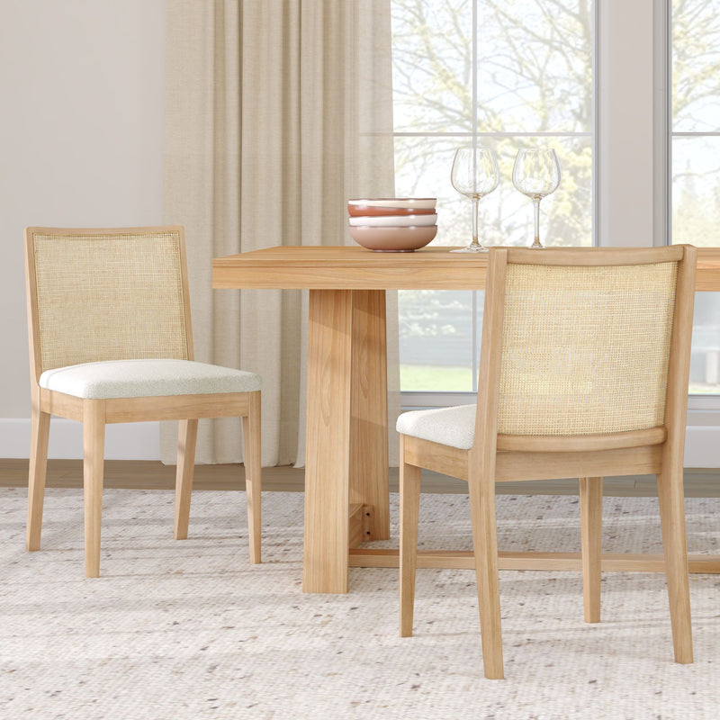 Light wood dining table with rattan cane-back chairs and upholstered white seats, ceramic bowls and wine glasses in a bright neutral dining room