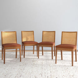 Four wooden dining chairs with woven backs and orange cushions on a beige carpet.