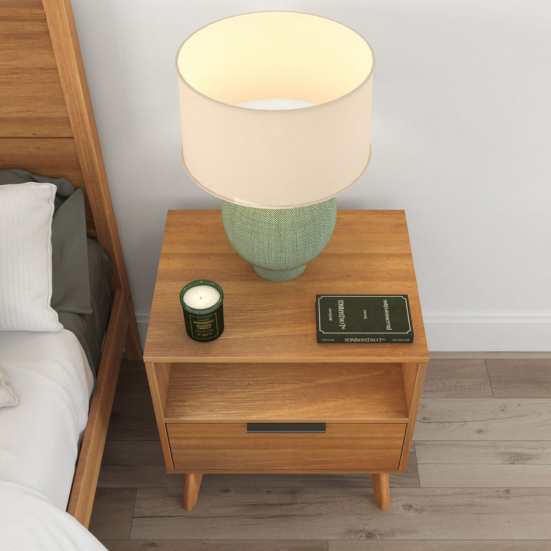 Wooden bedside table with modern lamp, candle, and book in cozy bedroom setting.