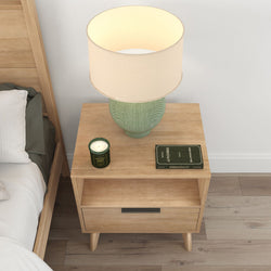 Wooden nightstand with a green table lamp, a lit candle, and a book in a cozy bedroom setting.