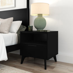 Black mid-century modern nightstand with drawer and open shelf, green textured ceramic lamp, candle and book on top, white bedding and light wood floor in minimalist bedroom
