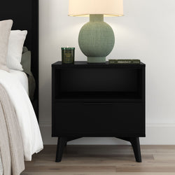 Black mid-century modern bedside table with open shelf and drawer, styled with a green textured table lamp, scented candle and book in a contemporary bedroom