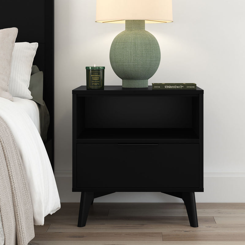 Black mid-century modern bedside table with open shelf and drawer, styled with a green textured table lamp, scented candle and book in a contemporary bedroom