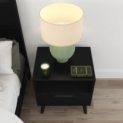Minimalist black nightstand with green ceramic table lamp, scented candle and hardcover book beside bed on light hardwood floor — modern bedroom bedside decor