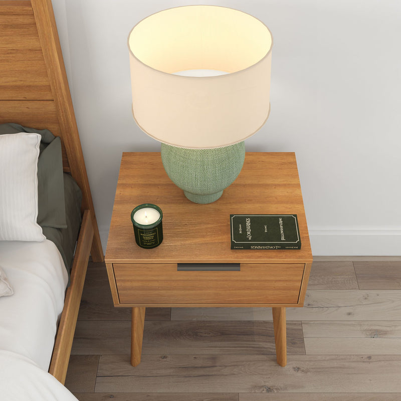 Wooden bedside table with drawer featuring a cream-colored lamp, green candle, and open book in a modern bedroom setting.