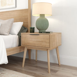 Wooden bedside table with drawer, featuring a modern table lamp, next to a neatly made bed in a cozy bedroom setting.