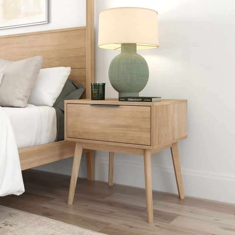 Wooden bedside table with drawer, featuring a modern table lamp, next to a neatly made bed in a cozy bedroom setting.
