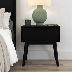 Modern black mid-century bedside table with drawer beside white bed, topped with textured green ceramic lamp, lit green scented candle and book on light wood floor — minimalist bedroom decor
