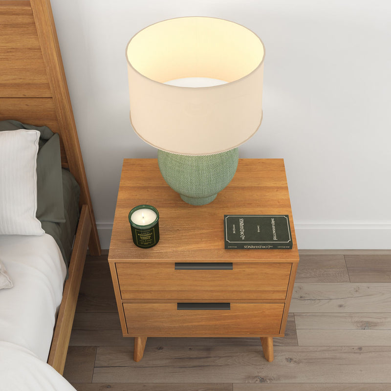 Wooden nightstand with lamp, candle, and book beside a bed in a cozy bedroom setting.