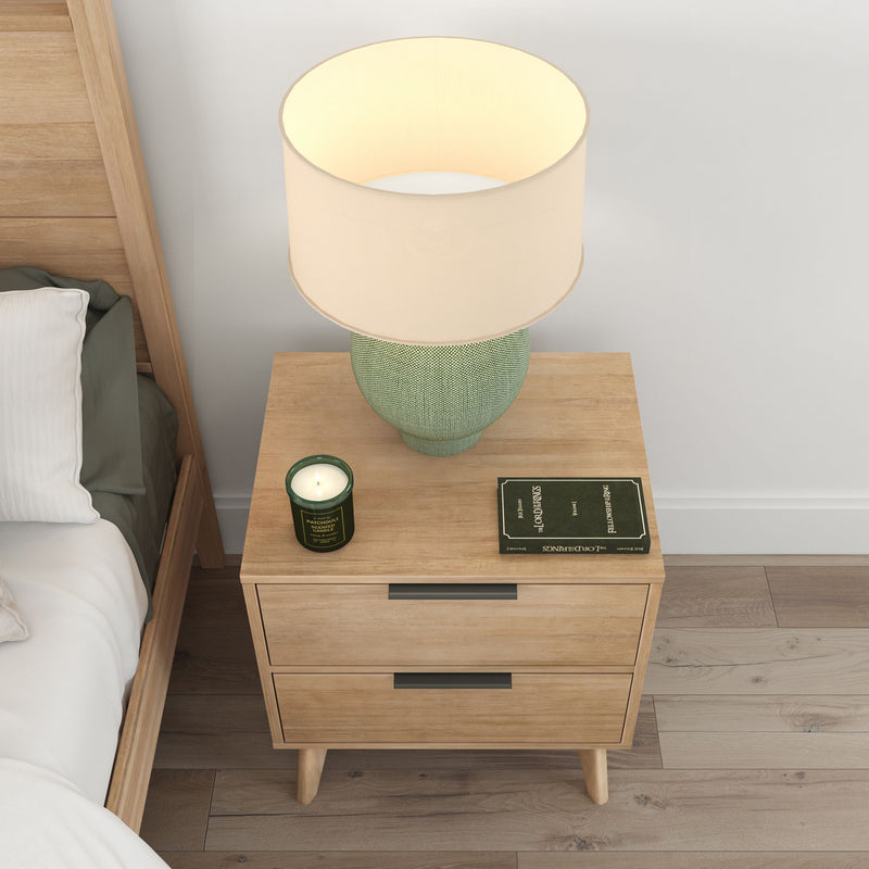 Wooden nightstand with green table lamp, scented candle, and book, in a cozy bedroom setting.