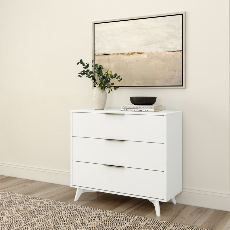 White three-drawer modern dresser with brass handles styled with a vase of greenery, black bowl and stacked books under neutral abstract wall art in a minimalist interior with wood floor and patterned rug
