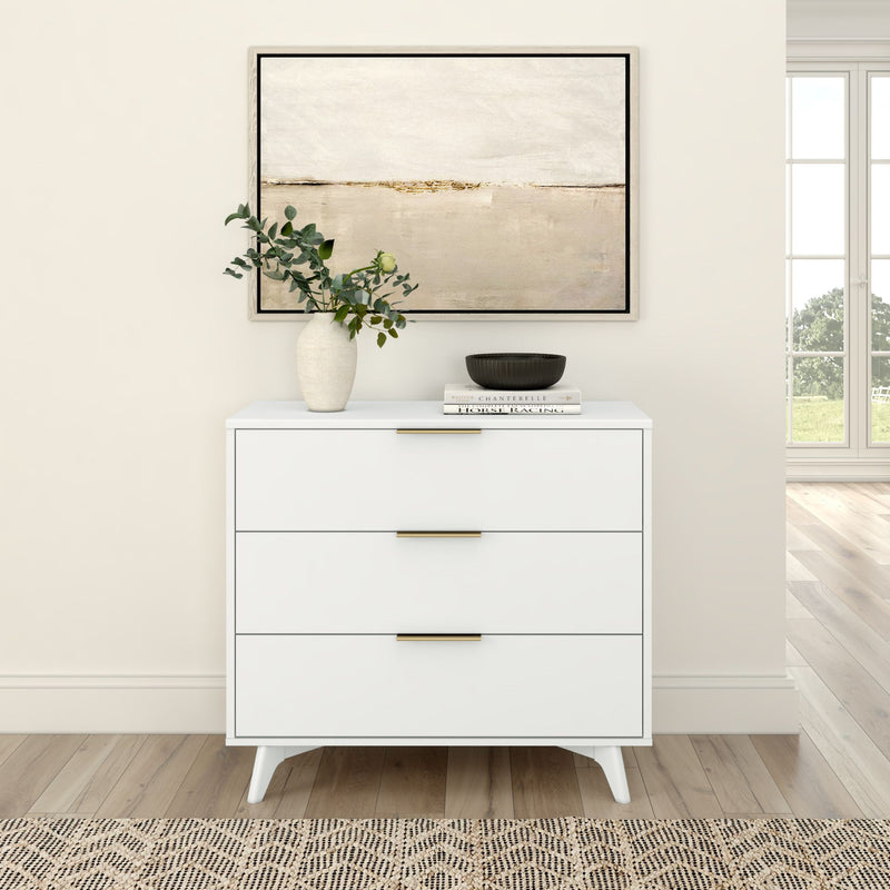 Modern white three-drawer dresser with brass handles, styled with vase of eucalyptus, books and abstract wall art in a sunlit minimalist interior on hardwood floors