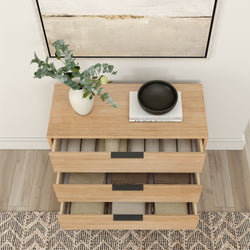 Wooden dresser with open drawers containing folded clothes, topped with a white vase of eucalyptus and a black bowl, placed on a hardwood floor against an abstract painting.