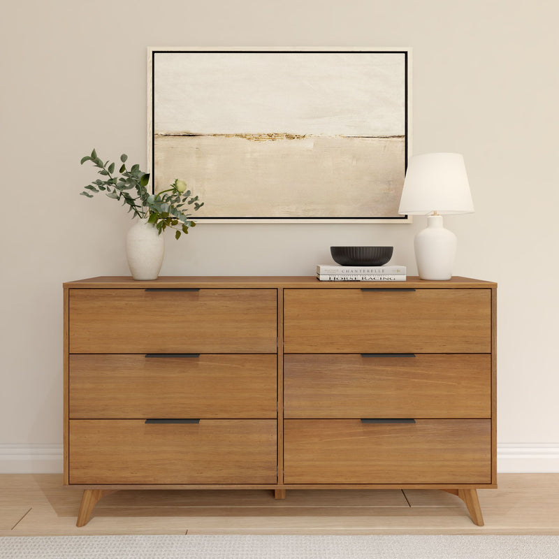 Mid-century modern six-drawer wooden dresser with white table lamp, vase of eucalyptus, stacked books and abstract wall art in neutral minimalist bedroom decor.