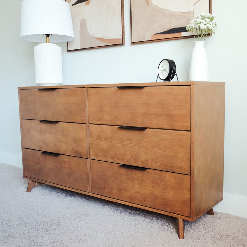 Mid-century modern six-drawer wooden dresser in warm walnut finish with black handles, styled with white table lamp, round black alarm clock and white vase of flowers in a bright carpeted bedroom