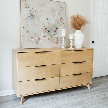 Light wood mid-century modern six-drawer dresser with black handles, styled with abstract wall art, white ceramic vase of dried flowers and white candlesticks on gray laminate flooring