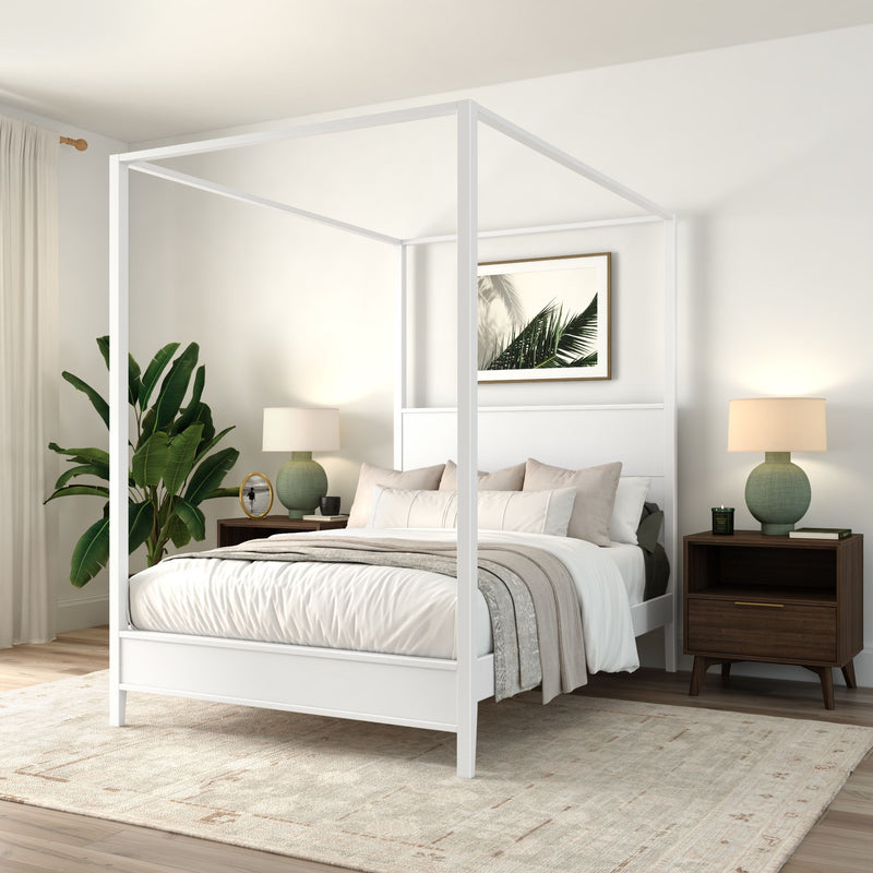 White four‑poster canopy bed in a modern neutral bedroom with layered beige bedding, mid-century wood nightstands, green ceramic lamps, large potted plant, palm print wall art and textured area rug