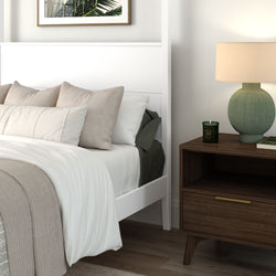 Modern minimalist bedroom with white bed frame, beige pillows, knit throw blanket, dark wood nightstand, green table lamp and scented candle