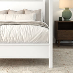 White four-poster bed with cream bedding and beige pillows, textured gray throw, wooden mid-century nightstand with green ceramic lamp and candle on a neutral area rug