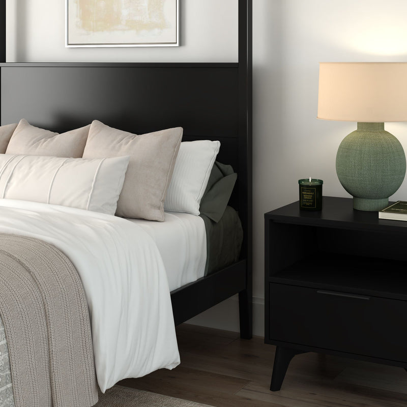 Cozy modern bedroom with black bed frame, white linens, beige throw and neutral pillows, black nightstand with green ceramic lamp, scented candle, and hardwood floor.