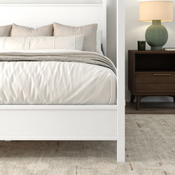 Minimalist modern bedroom with white platform bed and beige linen bedding, layered neutral throws and pillows, dark wood mid-century nightstand with green ceramic lamp and scented candle on a textured area rug