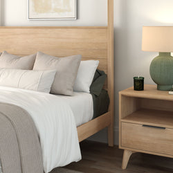 Modern minimalist bedroom interior with light wood bed frame and matching nightstand, white and beige bedding and decorative pillows, green ceramic table lamp and scented candle — cozy Scandinavian-style bedroom decor