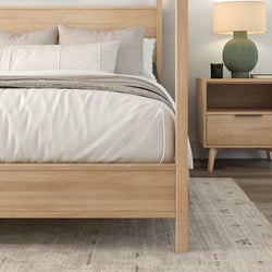 Modern Scandinavian light-wood four-poster bed with white bedding and beige pillows, matching nightstand with green lamp on neutral rug