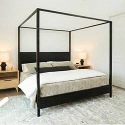 Modern black metal four-poster canopy bed in a neutral minimalist bedroom with beige quilted bedding, wooden nightstands, table lamps and patterned area rug