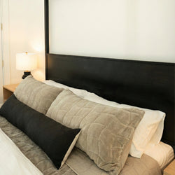 Modern bedroom decor featuring a black wooden headboard, neutral quilted bedding and beige pillows with a long black lumbar pillow, beside a wooden nightstand with a table lamp.