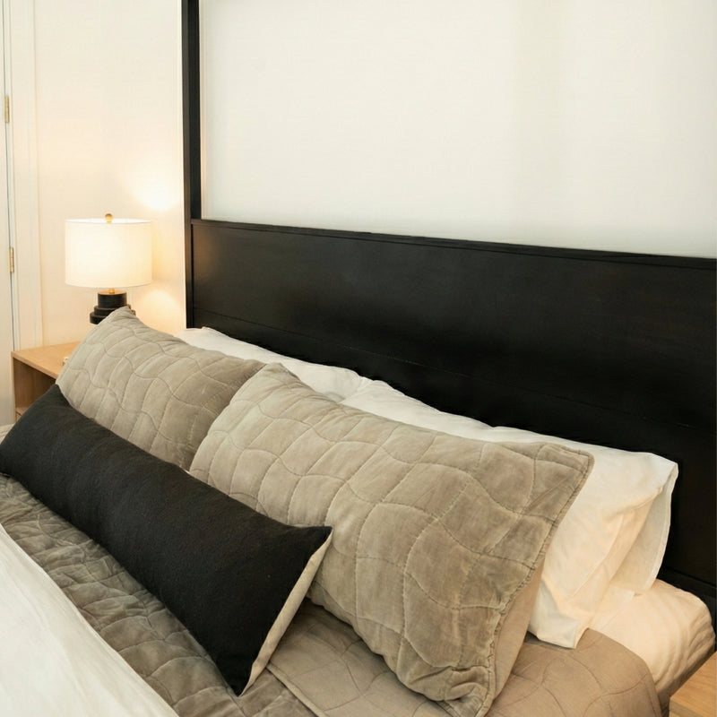 Modern bedroom decor featuring a black wooden headboard, neutral quilted bedding and beige pillows with a long black lumbar pillow, beside a wooden nightstand with a table lamp.