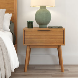 Mid-century modern wooden nightstand with drawer, featuring a green table lamp and scented candle, in a cozy bedroom setting.