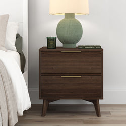 Mid-century modern dark walnut two-drawer nightstand with brass handles topped with a green textured table lamp, patchouli scented candle and book next to a white bed in a minimalist bedroom