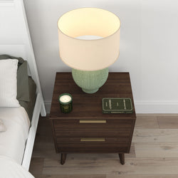 Mid-century modern dark wood nightstand with green ceramic table lamp and beige drum shade, scented candle and hardcover book beside a white bed on light hardwood floor