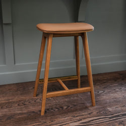 Mid-century modern wooden bar stool with tan leather cushion and brass footrest on dark hardwood floor against gray paneled wall