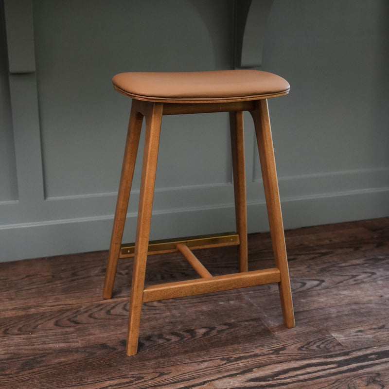 Mid-century modern wooden bar stool with tan leather cushion and brass footrest on dark hardwood floor against gray paneled wall