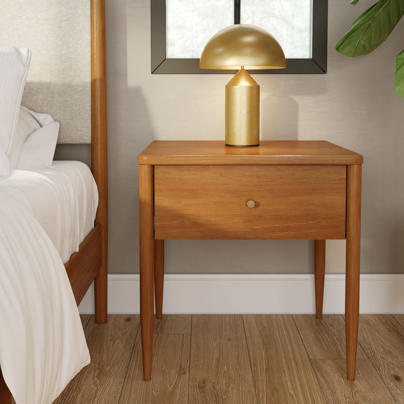 Mid-century modern wooden nightstand with single drawer and brass knob, gold mushroom table lamp on top, adjacent wooden bed frame with white bedding and hardwood floors in a cozy bedroom interior
