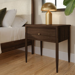 Mid-century modern dark wood bedside table nightstand with single drawer and brass knob, gold table lamp, white bedding and hardwood floor, stylish bedroom decor