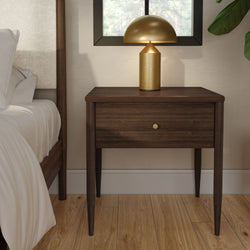 Mid-century modern dark wood nightstand with single drawer and gold table lamp beside bed on oak hardwood floor, minimalist bedroom interior