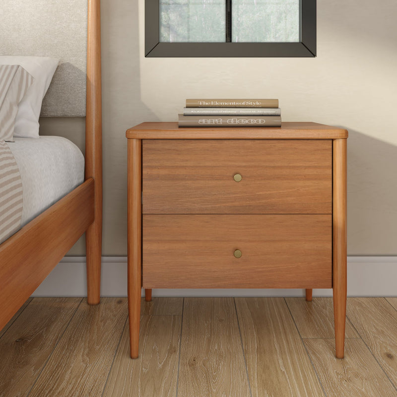 Mid-century modern wooden nightstand two-drawer bedside table with tapered legs and brass knobs in light oak finish, styled with stacked books beside a bed on hardwood floor for minimalist bedroom décor