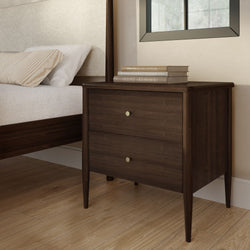 Dark walnut mid-century modern bedside table two-drawer nightstand with brass knobs beside upholstered bed on light hardwood floor with stacked books