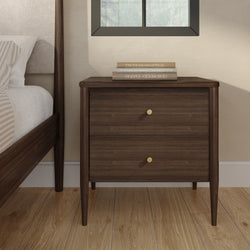 Walnut nightstand mid-century modern two-drawer bedside table with brass knobs next to upholstered bed on oak hardwood floor with stacked books and window in minimalist bedroom