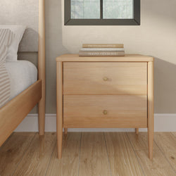 Light wood mid-century modern nightstand, two drawers with brass knobs, stacked books on top next to bed on hardwood floor