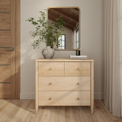 Minimalist light wood dresser with brass knobs, rectangular mirror, ceramic vase of greenery and stacked books in neutral modern bedroom decor