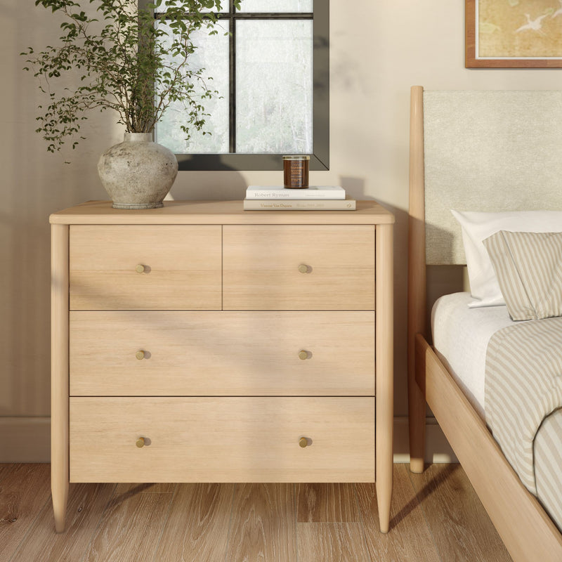 Light oak four-drawer bedroom dresser with brass knobs beside a minimalist upholstered bed, styled with a ceramic vase of greenery, stacked books and a candle for Scandinavian modern bedroom decor