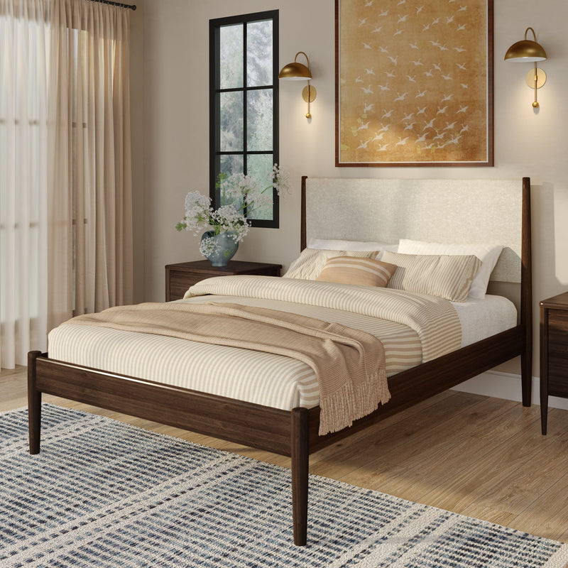 Mid-century modern bedroom with dark wood bed frame and linen headboard, beige striped bedding, brass wall sconces, wood nightstands, vase of white flowers, black-frame window and blue patterned rug