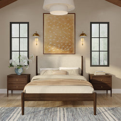 Modern neutral bedroom with mid-century wooden bed frame, beige striped bedding and throw, matching walnut nightstands, brass wall sconces, large framed bird artwork, tall black-framed windows, floral vase on left nightstand, and patterned area rug