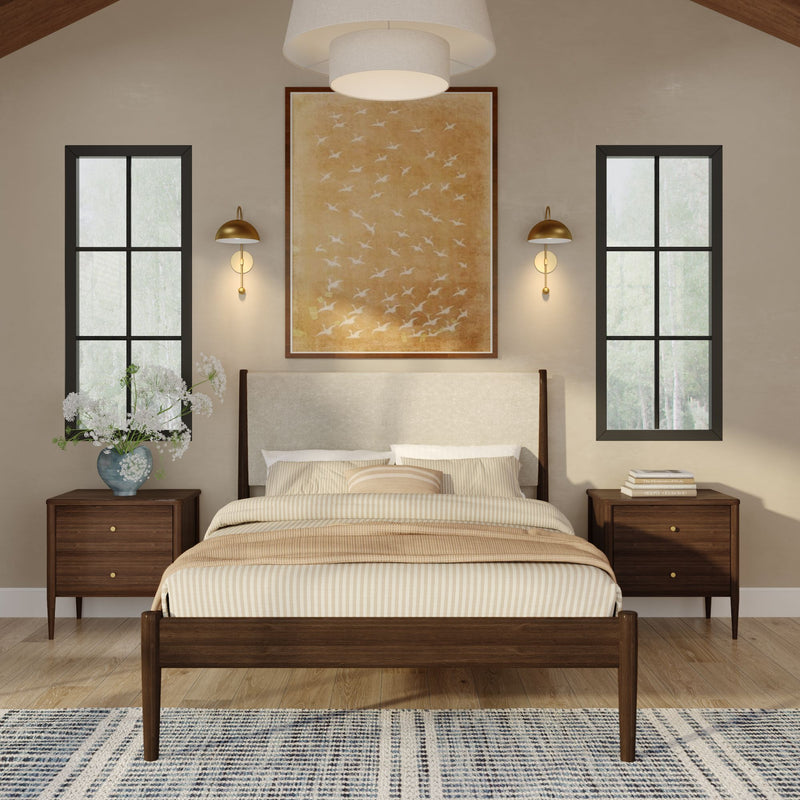 Modern neutral bedroom with mid-century wooden bed frame, beige striped bedding and throw, matching walnut nightstands, brass wall sconces, large framed bird artwork, tall black-framed windows, floral vase on left nightstand, and patterned area rug