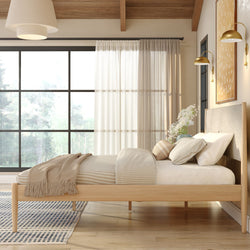 Sunlit modern Scandinavian bedroom with wooden platform bed, neutral linens and throw, large grid window with sheer curtains, gold wall sconces and minimalist decor