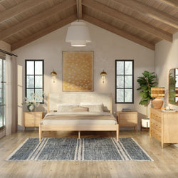 Modern farmhouse bedroom interior with vaulted wood ceiling, natural wood platform bed and nightstands, neutral linen bedding, brass wall sconces, black-framed windows, blue patterned rug, dresser with rattan lamp and fiddle leaf fig plant