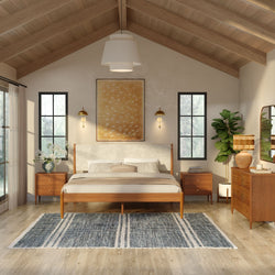 Mid-century modern bedroom with wooden platform bed, vaulted wood-beamed ceiling, large windows, brass wall sconces, matching nightstands, woven area rug and potted plant in a neutral, natural-light interior